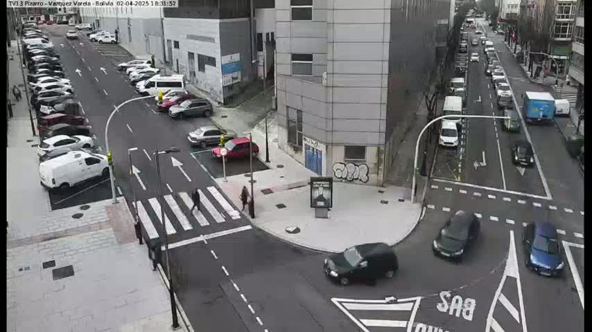 A street intersection with parked and moving cars, pedestrians crossing a crosswalk, and a multi-story building in the background on an overcast day.