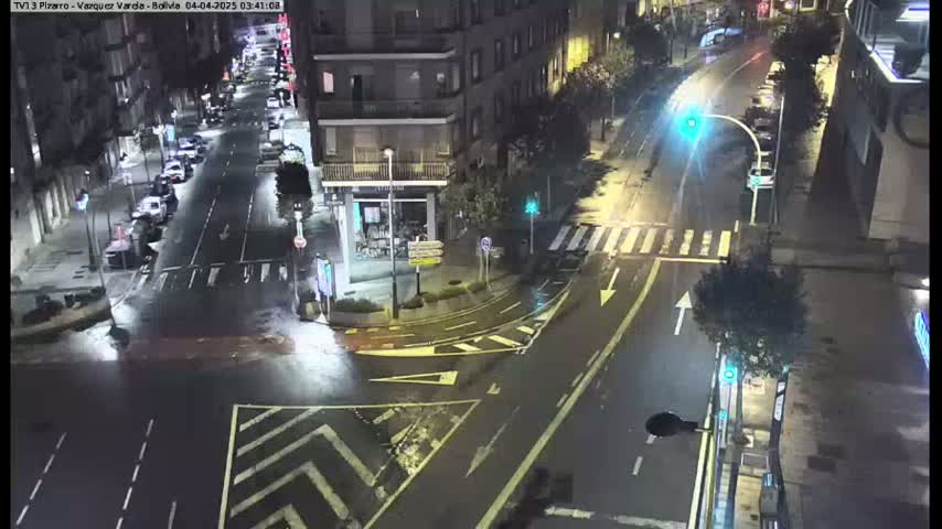 A wet, nighttime street intersection shows several cars parked and driving, with buildings visible in the background.