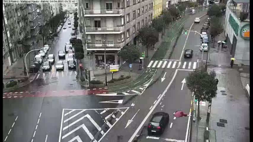 A rainy city intersection shows several cars navigating a wet roadway and crosswalks, with buildings and trees lining the streets.