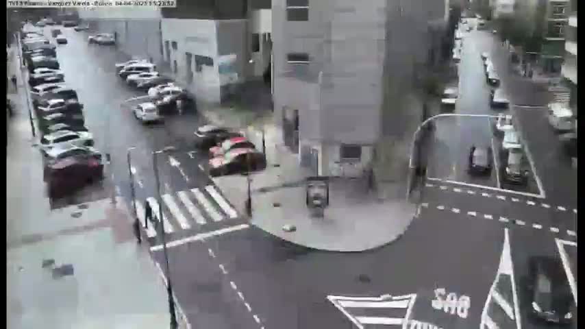 A rainy city street intersection shows several parked cars along the sides of the street and several cars moving through the intersection.