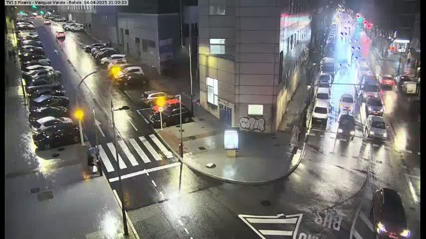 A rain-slicked city street at night shows several cars parked along the side and others driving through a busy intersection.