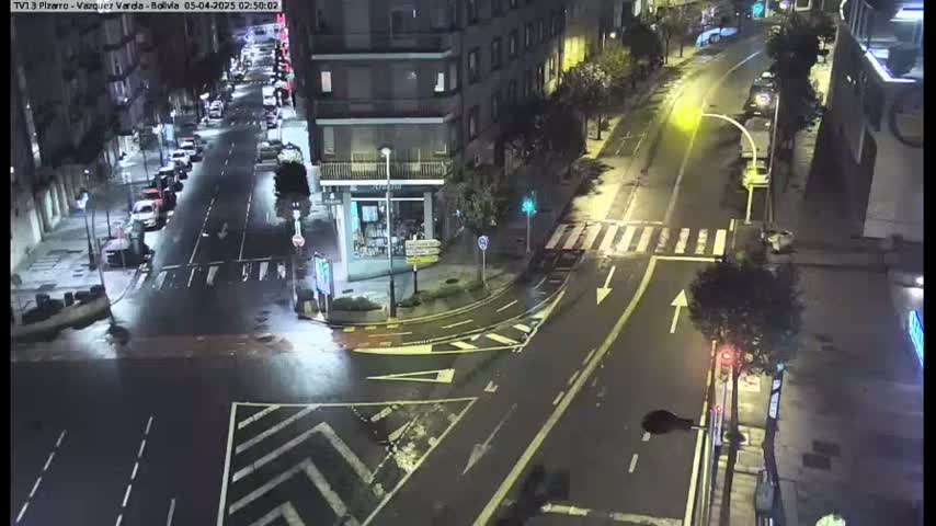 A wet, nighttime street intersection in a city, with cars parked along the sides of the road and a few cars driving through.