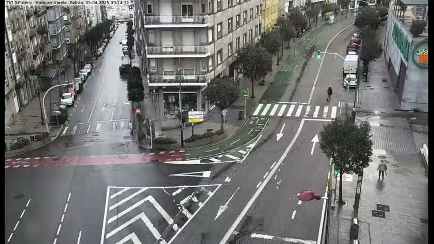 A wet city street intersection with parked cars and pedestrians, showing a bike lane and several buildings in the background.