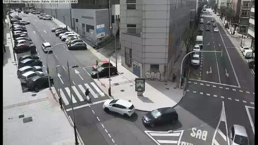 A sunny city street intersection shows several cars parked along the sides of the road and others driving through the intersection.