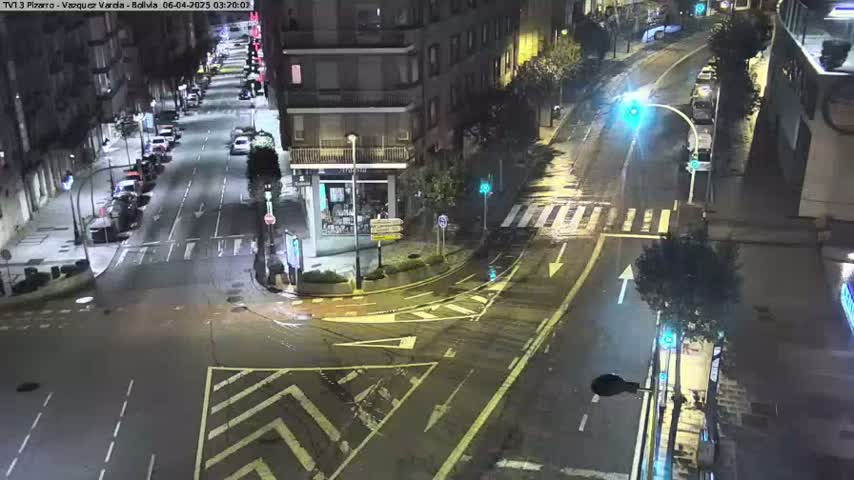 A wet, nighttime street intersection shows several cars parked along the sides of the roads and a few cars driving.