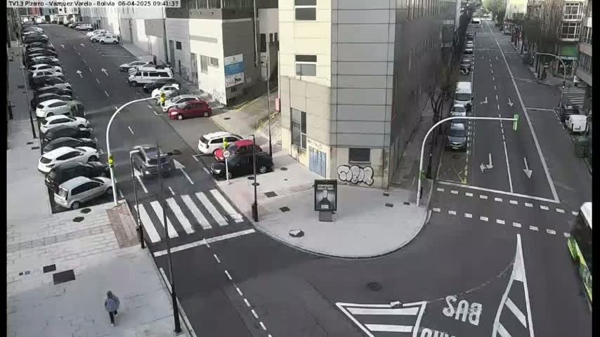 A street intersection with parked cars along the sides, a pedestrian crossing, and a few cars driving on a sunny day.
