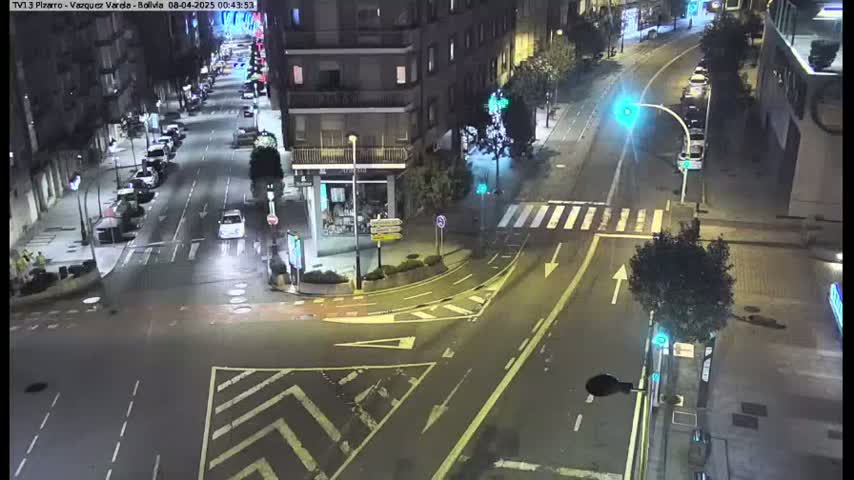 A nighttime, high-angle view of a city intersection with several cars parked along the sides of the streets and a few cars driving through the intersection.