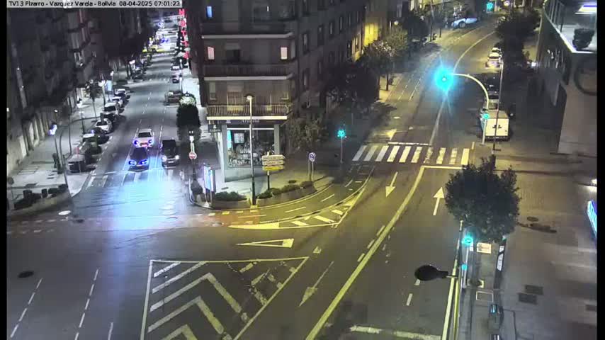 A nighttime street intersection with several cars and a van, showing city buildings and some pedestrian crossings under slightly wet conditions.