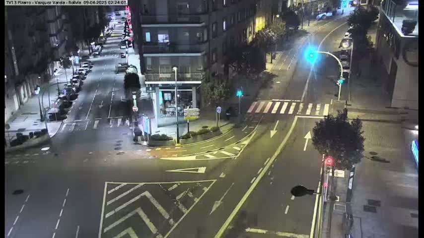 A nighttime, wet city street intersection with sparsely parked cars along the sides and a few vehicles at the crossing.