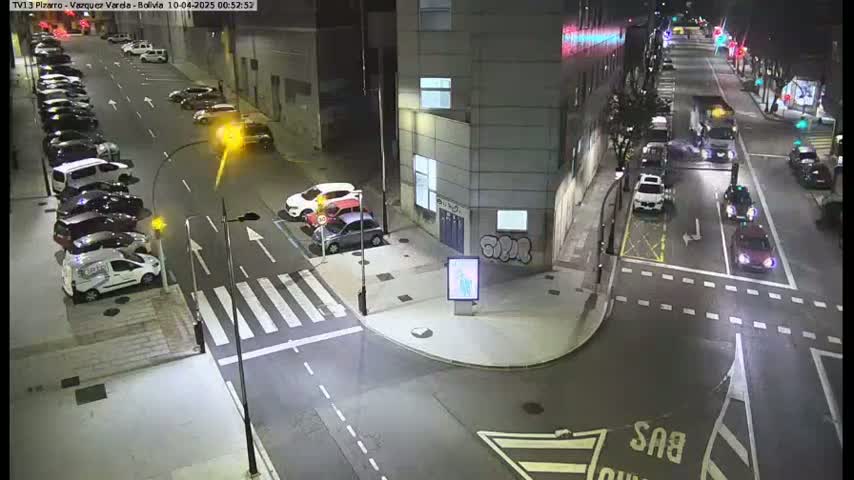 A nighttime aerial view of a city intersection with parked cars lining the streets and several vehicles moving through the crossroads.