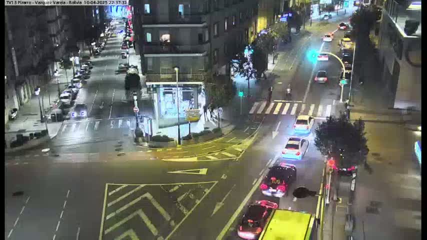 A nighttime aerial view of a city street intersection with several cars at a traffic light and buildings lining the street.