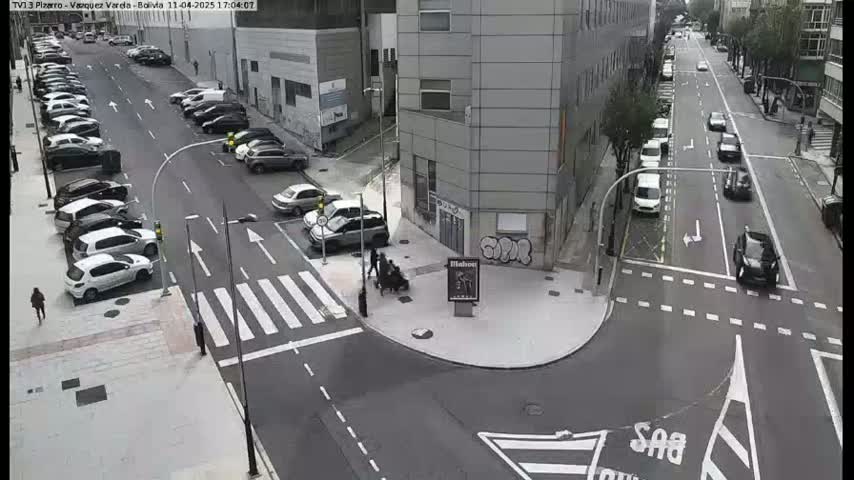 A city street intersection with parked cars along the sides and a few cars driving through, under an overcast sky.