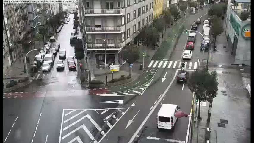 A wet city intersection with multiple cars, a white van, and a person on a scooter, surrounded by buildings on a rainy day.