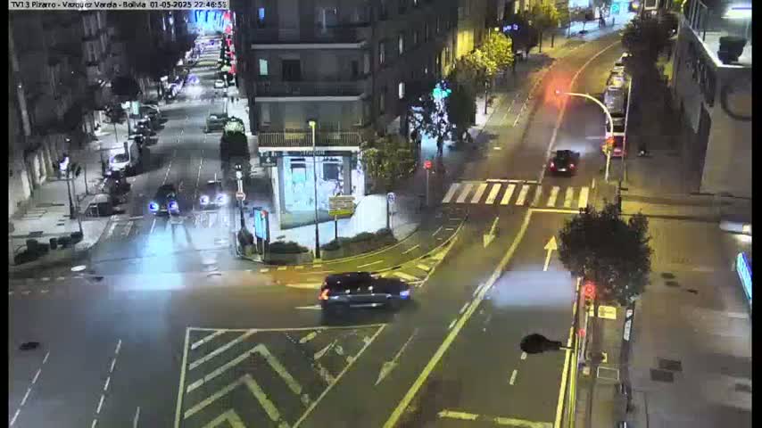 A nighttime, wet street intersection shows several cars driving on roadways, with buildings and streetlights visible in the background.