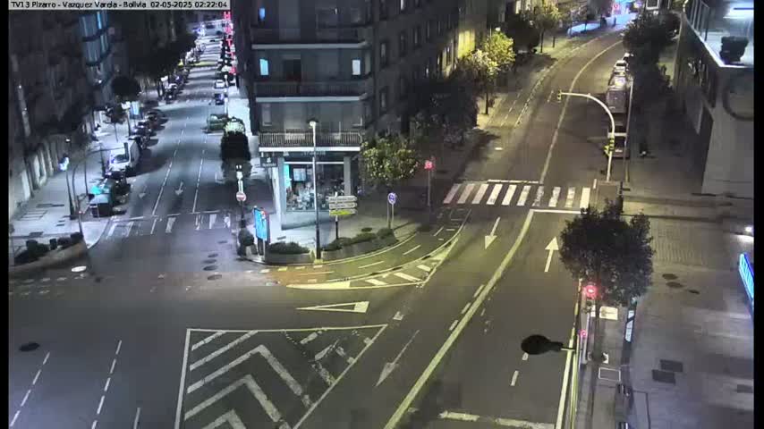 A nighttime, street-level view of a mostly empty intersection with cars parked along the sides of the roads and buildings lining the streets.