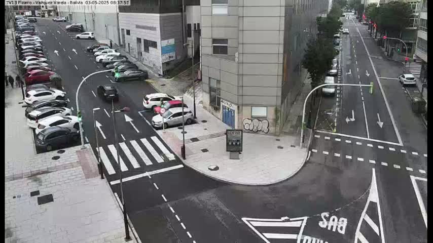 A wet city street intersection shows cars parked along the sides of the street and a few cars moving through the intersection.