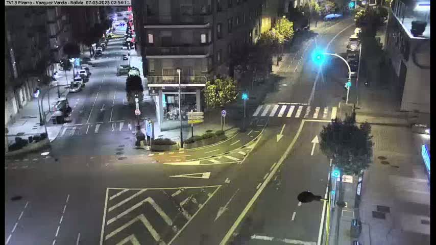 A nighttime aerial view of a city intersection with several cars parked along the street and a few cars driving, under well-lit conditions.
