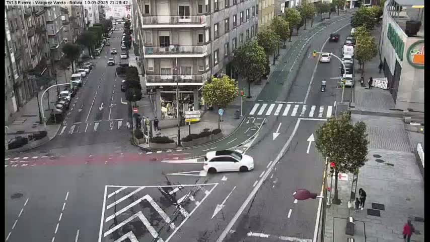A street intersection in a city, showing several cars and pedestrians, on a partly cloudy day.