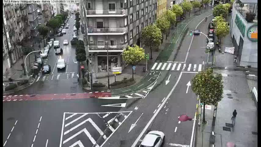 A wet city street intersection shows several cars at a crosswalk, with buildings and trees lining the streets on a rainy day.