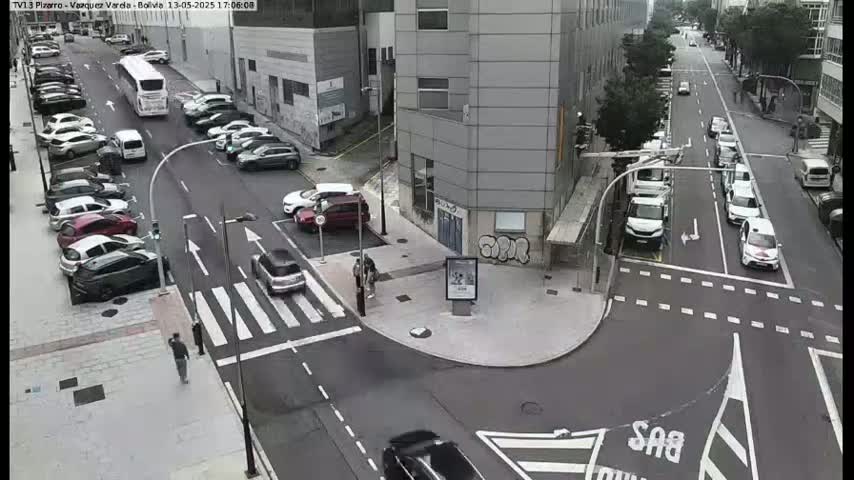 A street intersection with parked cars along the sides and a few cars driving through, on a partly cloudy day.