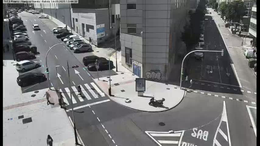 A sunny city street scene shows parked cars lining both sides of a road that intersects with another road, where a motor scooter is parked near a building and pedestrians cross a crosswalk.