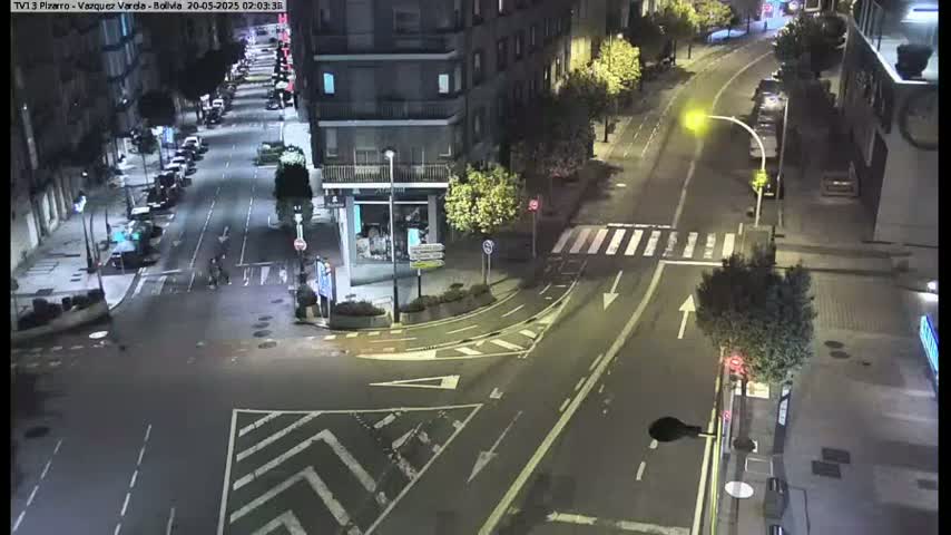 A nighttime, high-angle view of a city intersection with parked cars lining the streets, a few pedestrians, and streetlights illuminating the scene.