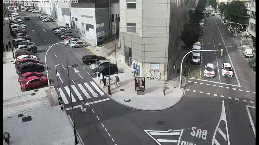A sunny city street intersection with parked cars along the sides and a few pedestrians near a building, with two taxis waiting at a traffic light.