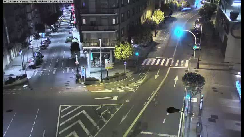 A wet nighttime street intersection shows several cars parked along the sides of the road, with a few more driving through the intersection under streetlights.