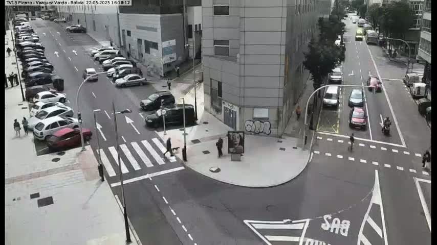 A city intersection with parked cars along the sides of the streets and several cars driving through, with pedestrians crossing a crosswalk, and a person walking a dog.