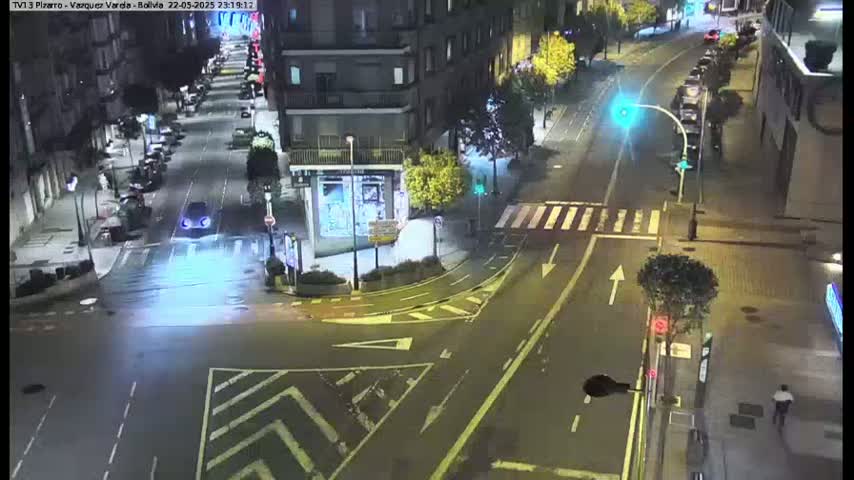A nighttime street intersection with parked cars along the sides of the road, wet pavement, and a few pedestrians.