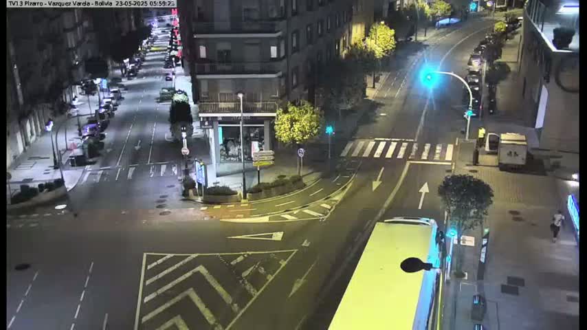A wet, nighttime street intersection in a city shows several cars parked along the sides of the roads, with a few cars driving through the intersection.