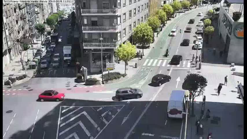 A sunny city street scene shows several cars at an intersection, with pedestrians walking on sidewalks and a bike lane visible.
