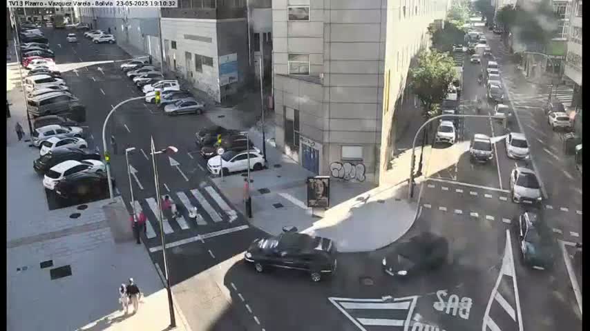 A sunny city street intersection shows several cars parked along the sides and several more driving through the intersection, with pedestrians crossing a crosswalk.