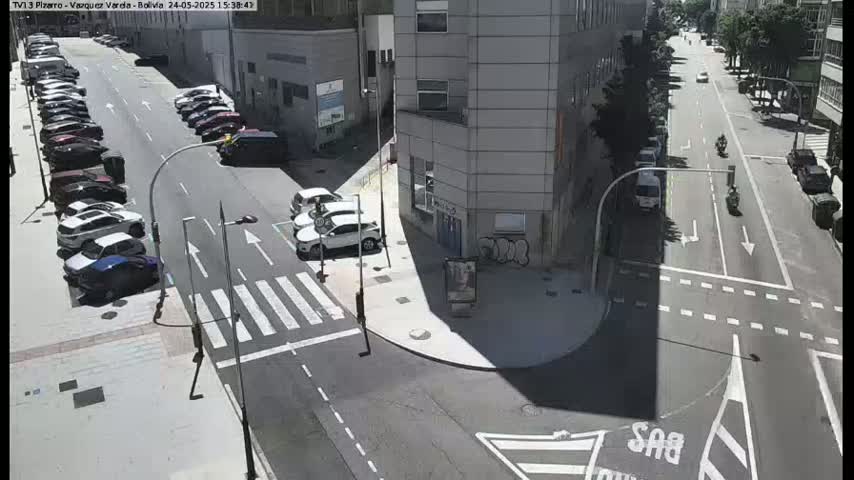 A sunny city street intersection with parked cars along the side of the road and several vehicles traveling through the intersection.