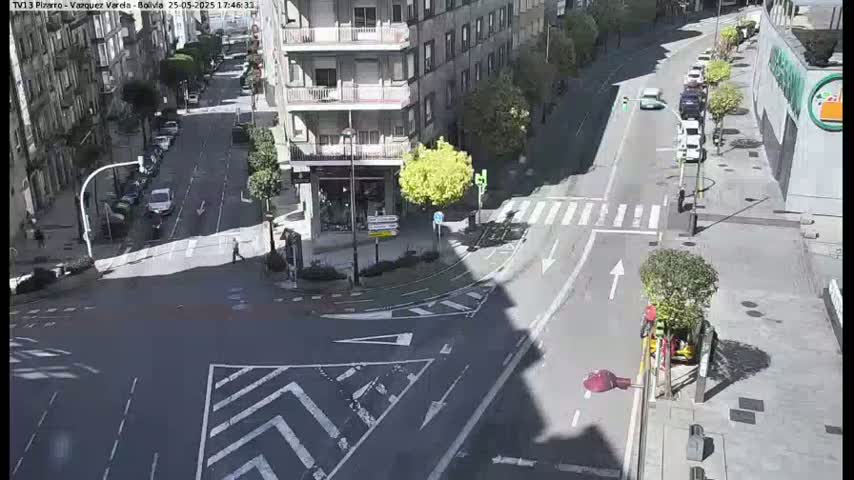 A sunny street intersection shows cars parked along the sides of the street and a few cars driving through the intersection.