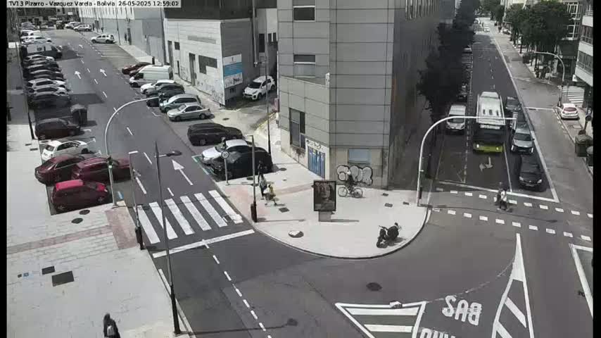A sunny street intersection shows several parked cars and a few moving vehicles, with pedestrians and cyclists visible.