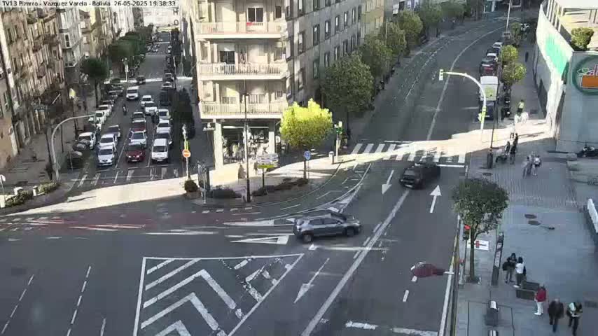 A sunny city street intersection shows several cars driving and pedestrians walking, with buildings lining the street.