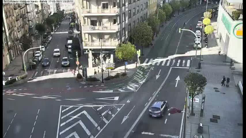 A sunny day shows a city intersection with several cars and pedestrians, and buildings lining the streets.