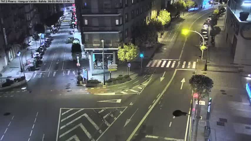 A nighttime view of a city intersection with parked cars along the sides of the street and sparsely trafficked roads.