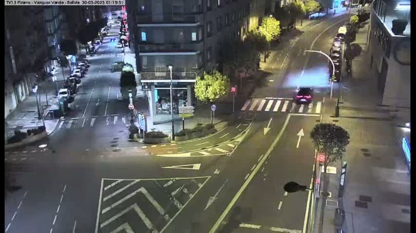 A nighttime aerial view shows a street intersection with several cars and buildings, wet pavement indicating recent rain.