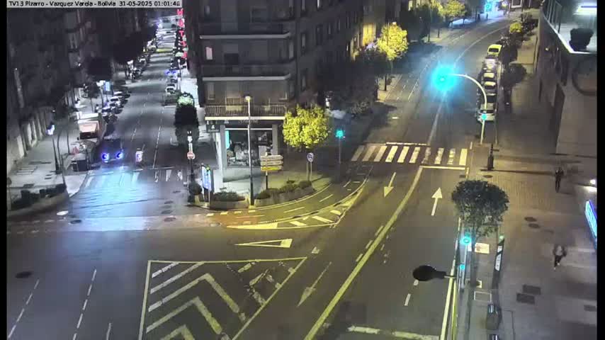 A nighttime, wet street intersection shows several cars, buildings, and pedestrians under streetlights.