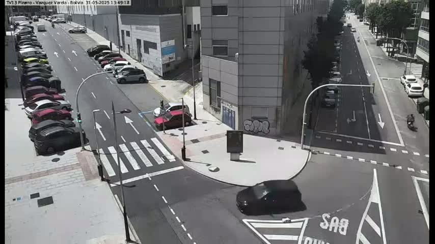 A sunny day shows a street intersection with cars parked along the sides of the road and a few cars driving through.