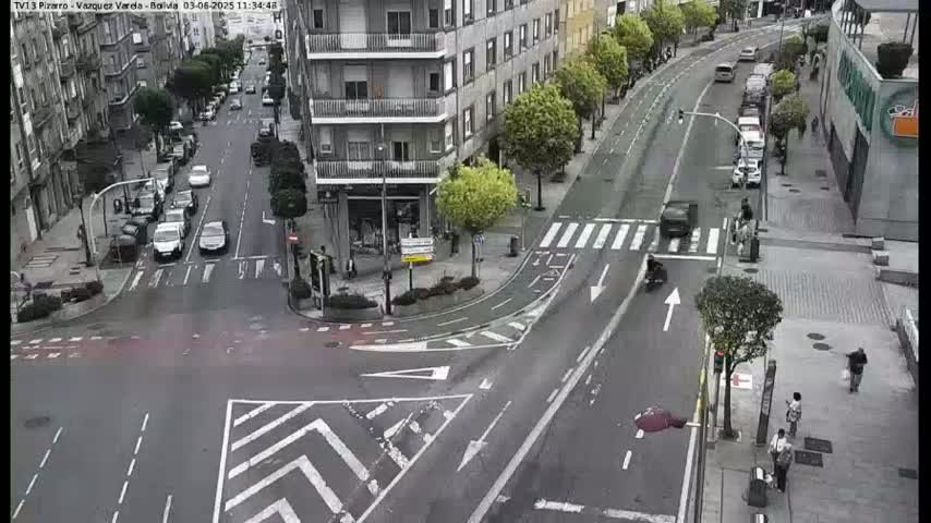 A city street intersection with several cars and pedestrians, under an overcast sky.