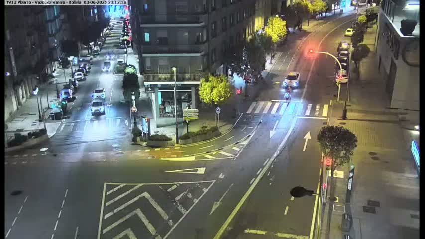 A nighttime, wet street intersection shows several cars, buildings, and trees under streetlights.
