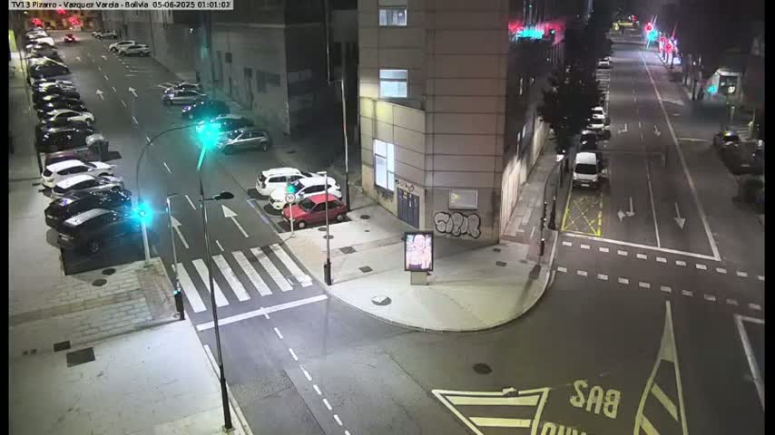 A nighttime, street-level view of a city intersection with parked cars along the sides of the road and a few cars driving by.