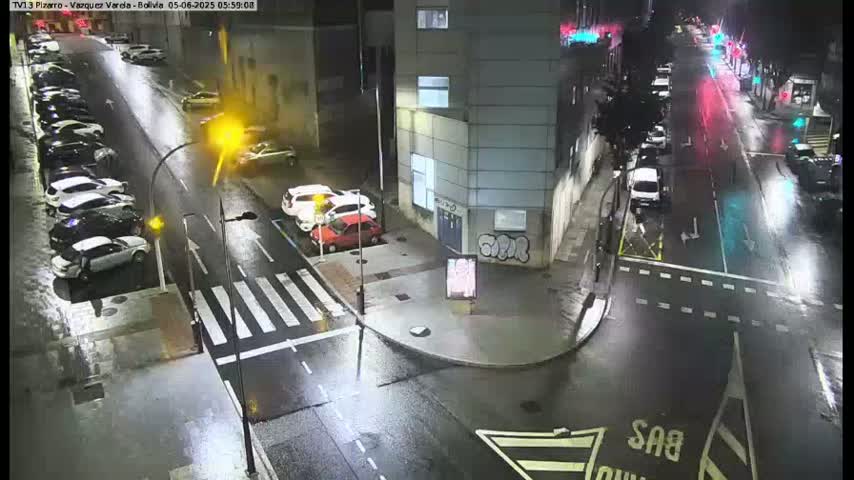 A wet, nighttime street scene shows several cars parked along the side of a road and others driving past a building.