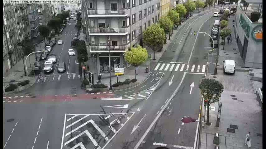 A wet city street intersection shows several cars and pedestrians, with buildings and trees lining the road.
