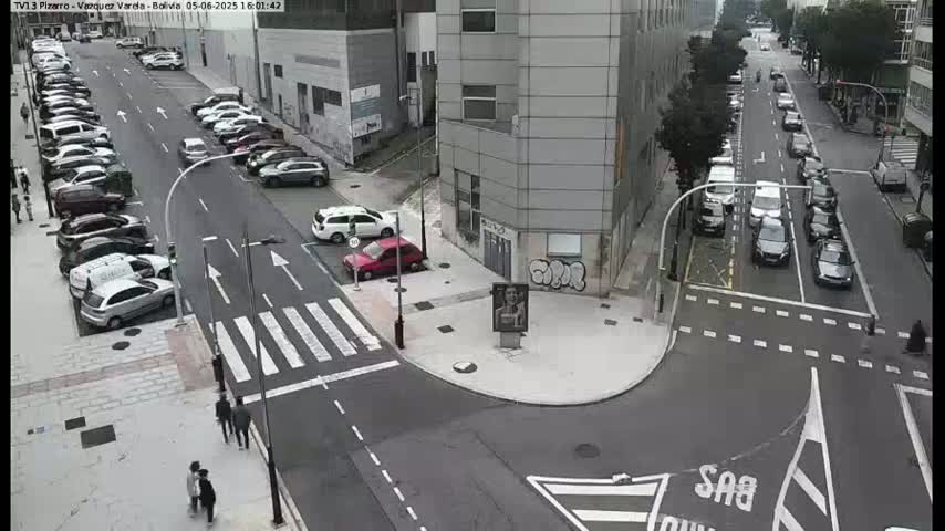 A high-angle view of a city intersection with several cars parked along the sides of the street and a few cars driving through, on a sunny day.
