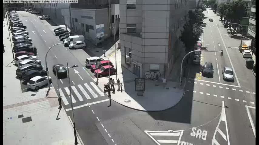 A sunny city street scene shows several cars parked along the side of the road and others driving through an intersection near a tall building.