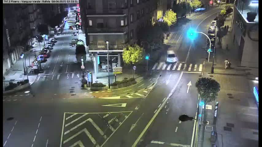 A nighttime, high-angle view of a city intersection with several cars and pedestrians, showing a street with parked vehicles along the sides and crosswalks.